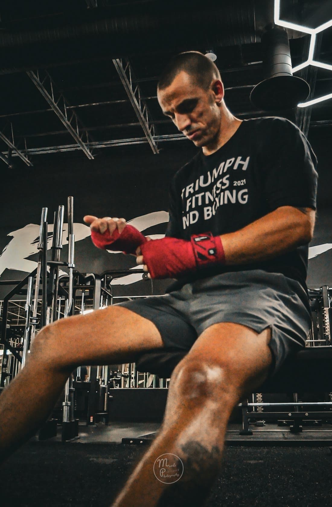 Eric Lawes seated in a dark gym wrapping red hand wraps, wearing a Triumph Fitness and Boxing shirt under hexagonal LED lights