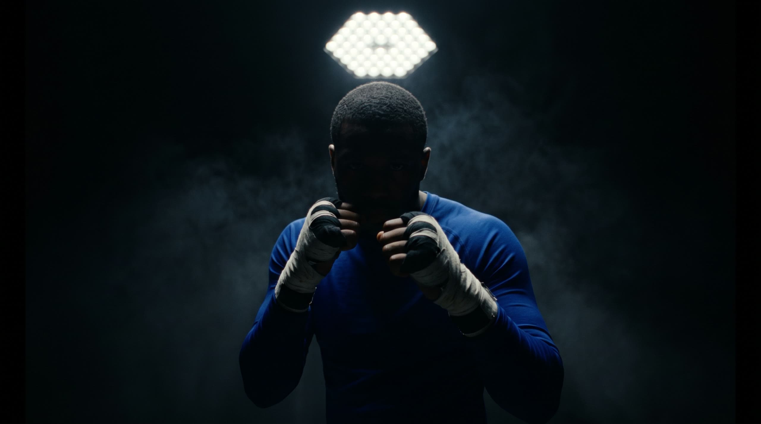 Boxer in guard stance with wrapped fists raised, rim-lit by a hexagonal LED cluster